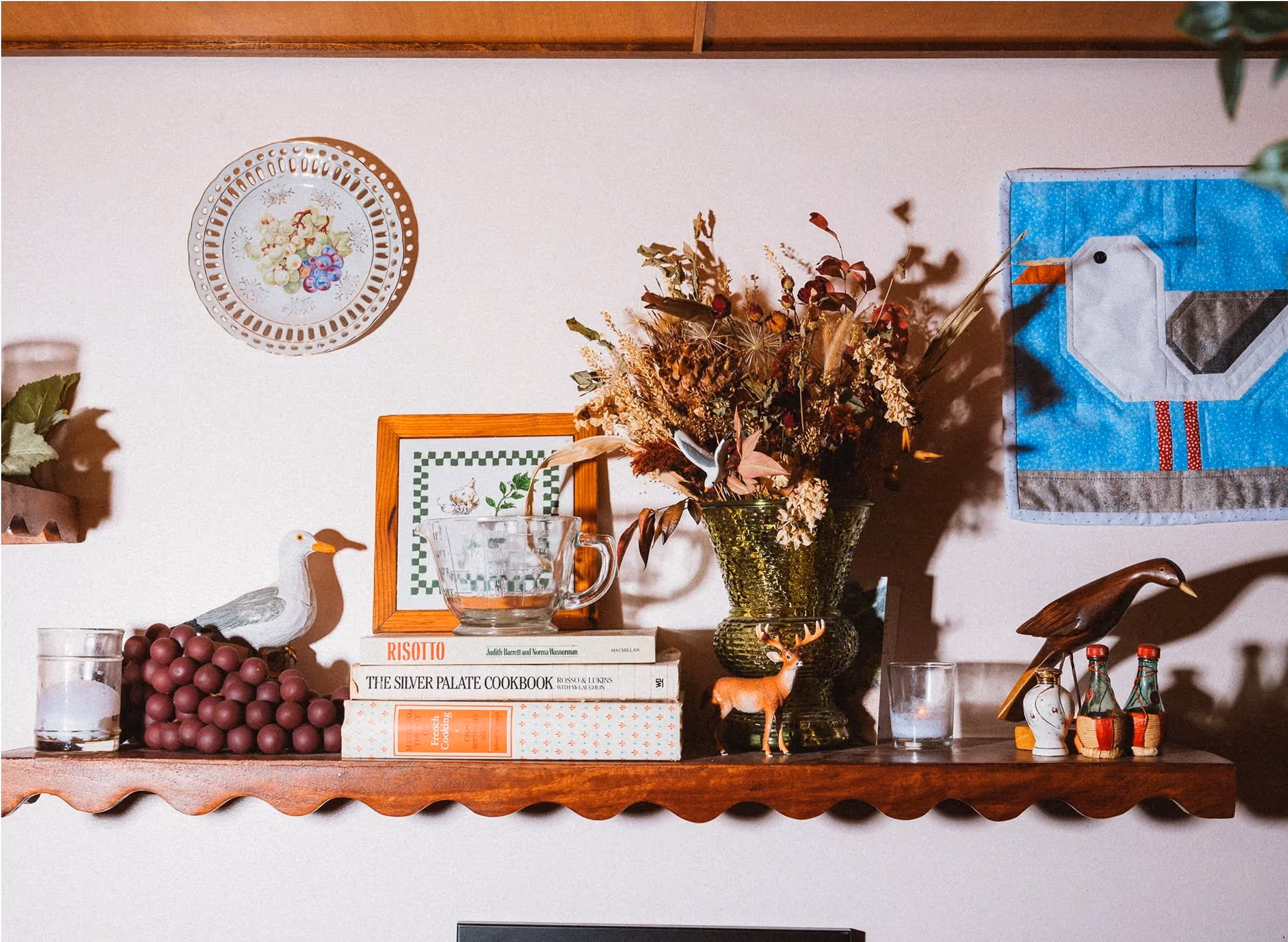 A wooden shelf displays books, a vase with dried flowers, bird figurines, candles, and a stack of chocolate balls. Decorative art featuring birds hangs on the wall behind the shelf.