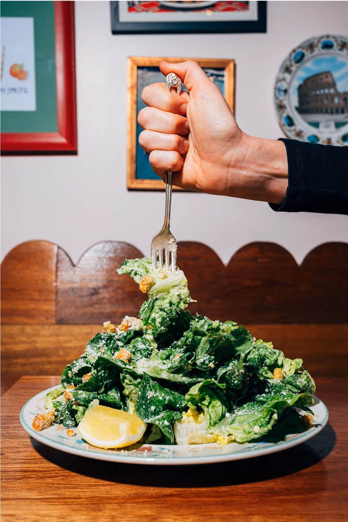 A hand holding a fork vertically above a large plate of Caesar salad with leafy greens, croutons, and lemon wedges on a wooden table, with framed art on the wall in the background.