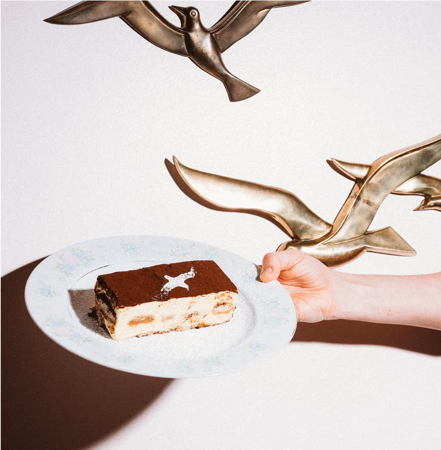 A hand holds a plate with a slice of tiramisu; in the background, there are metallic bird wall decorations casting shadows on a white wall.