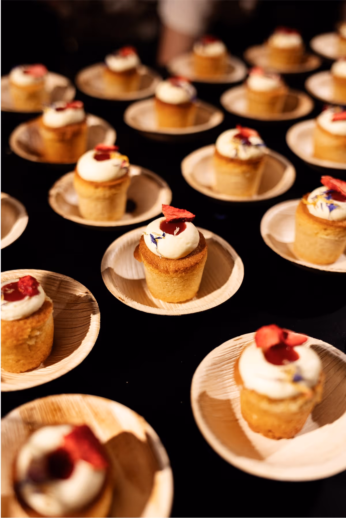 Rows of small cupcakes in wooden dishes, each topped with white icing and a garnish of dried red fruit, displayed on a dark surface.