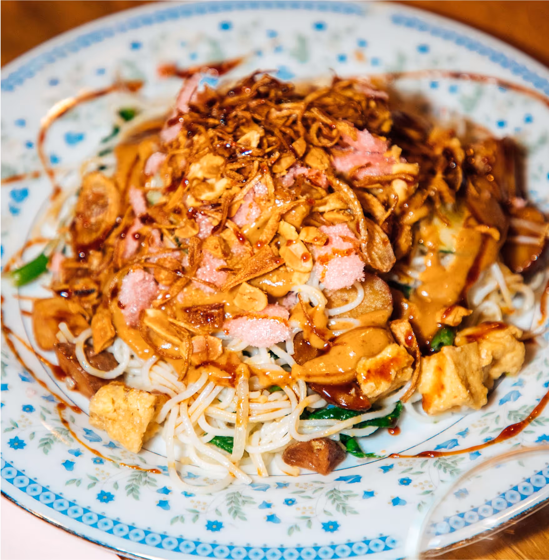 A plate of Indonesian-style noodles topped with peanut sauce, crispy fried shallots, tofu cubes, and a drizzle of sweet soy sauce on a decorative white and blue plate.