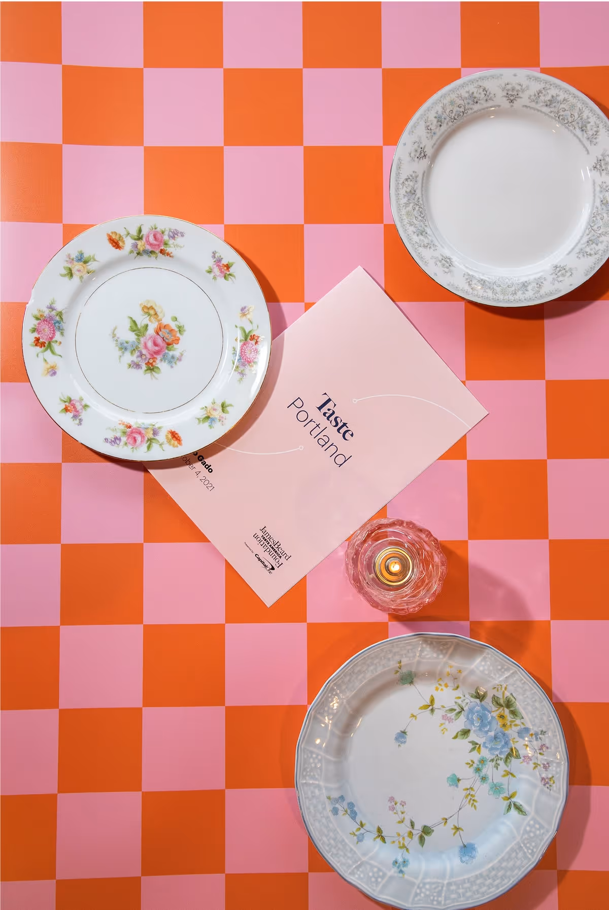 Three vintage-style plates surround a pink event program titled Taste Portland on a pink and orange checkered tablecloth, with a small lit candle in a glass holder nearby.