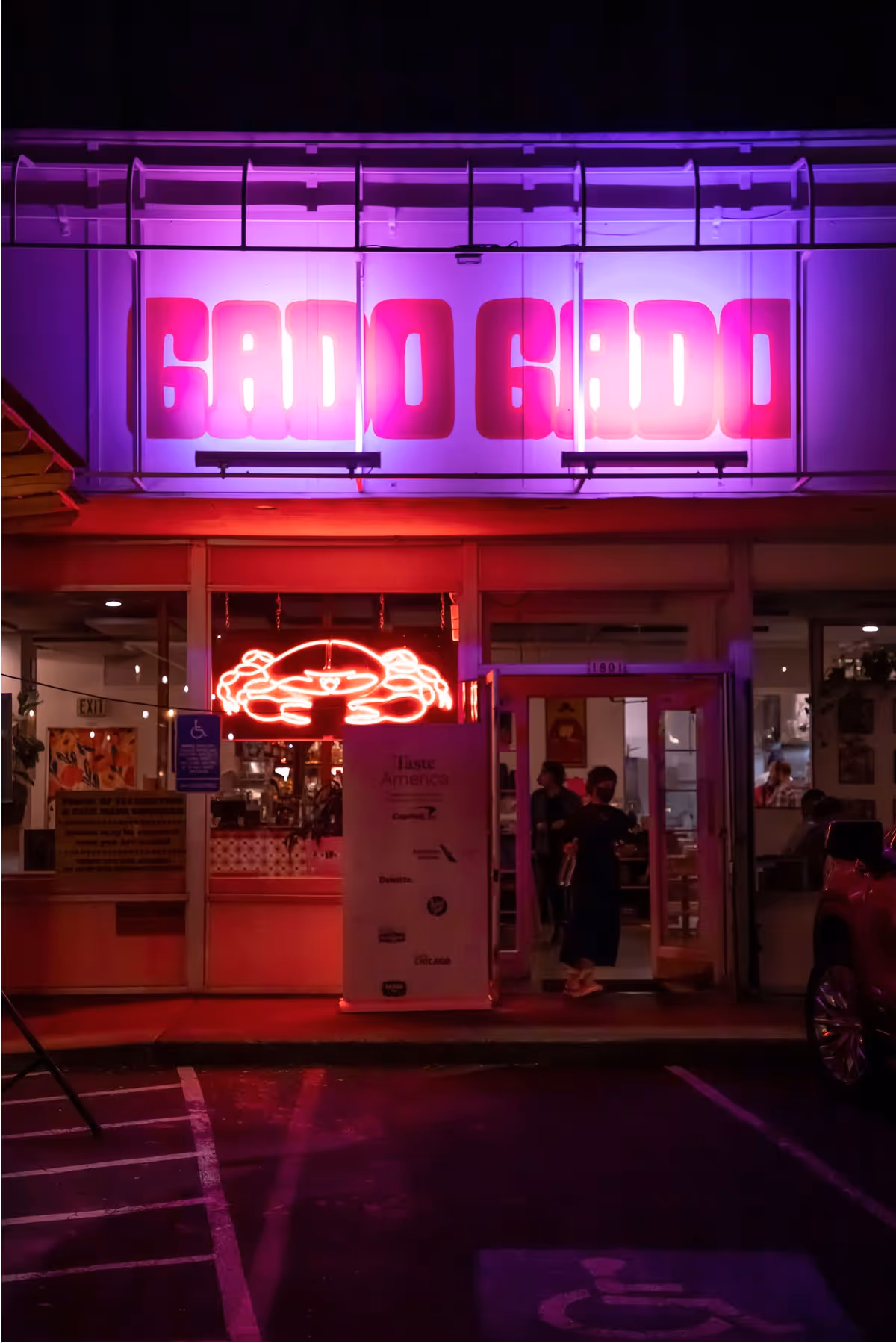 A restaurant entrance at night with bright pink neon signs, including GADO GADO above the door and a glowing crab below. Two people stand inside near the glass entrance, and a red car is parked outside.