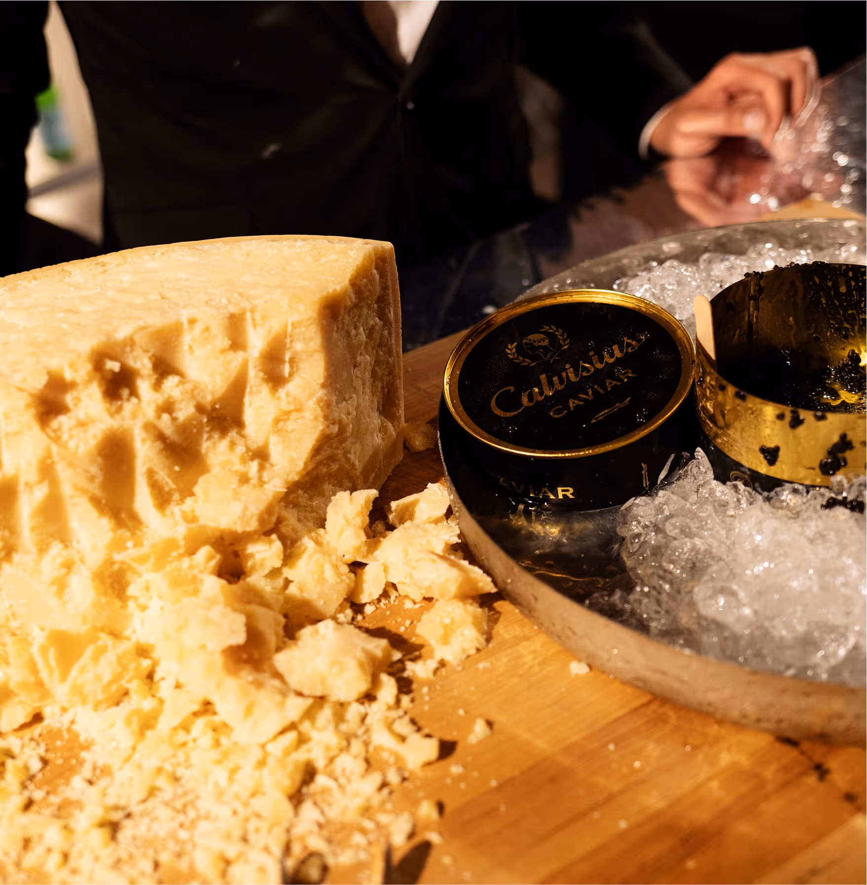 A large wedge of crumbled cheese sits on a wooden board next to an open tin of Calvisius caviar on ice. A person in a black suit stands behind the table.