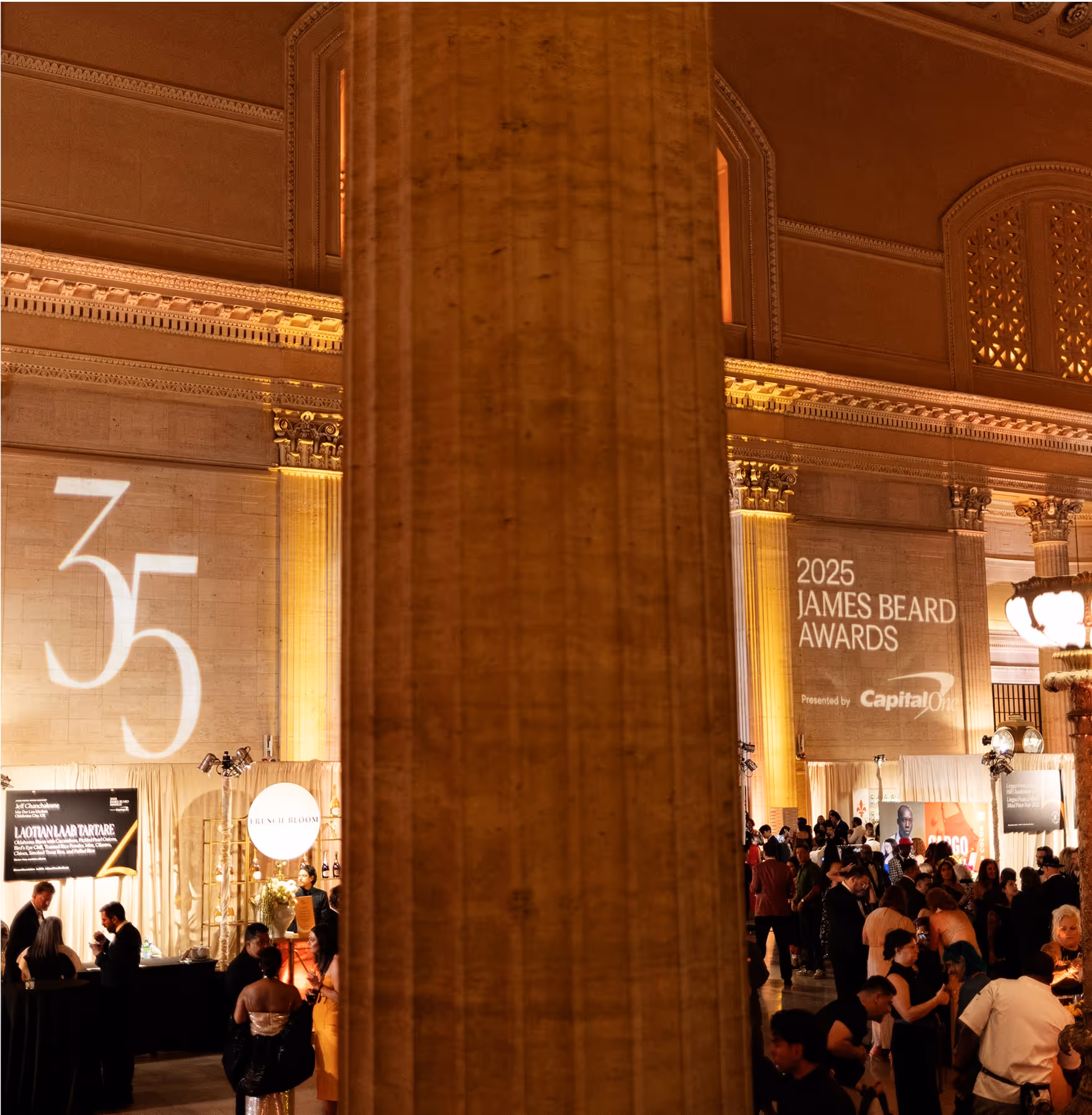 A large pillar stands in the center of a grand hall filled with people attending the 2025 James Beard Awards. Light projections display “35” and event details on the walls above food and sponsor booths.