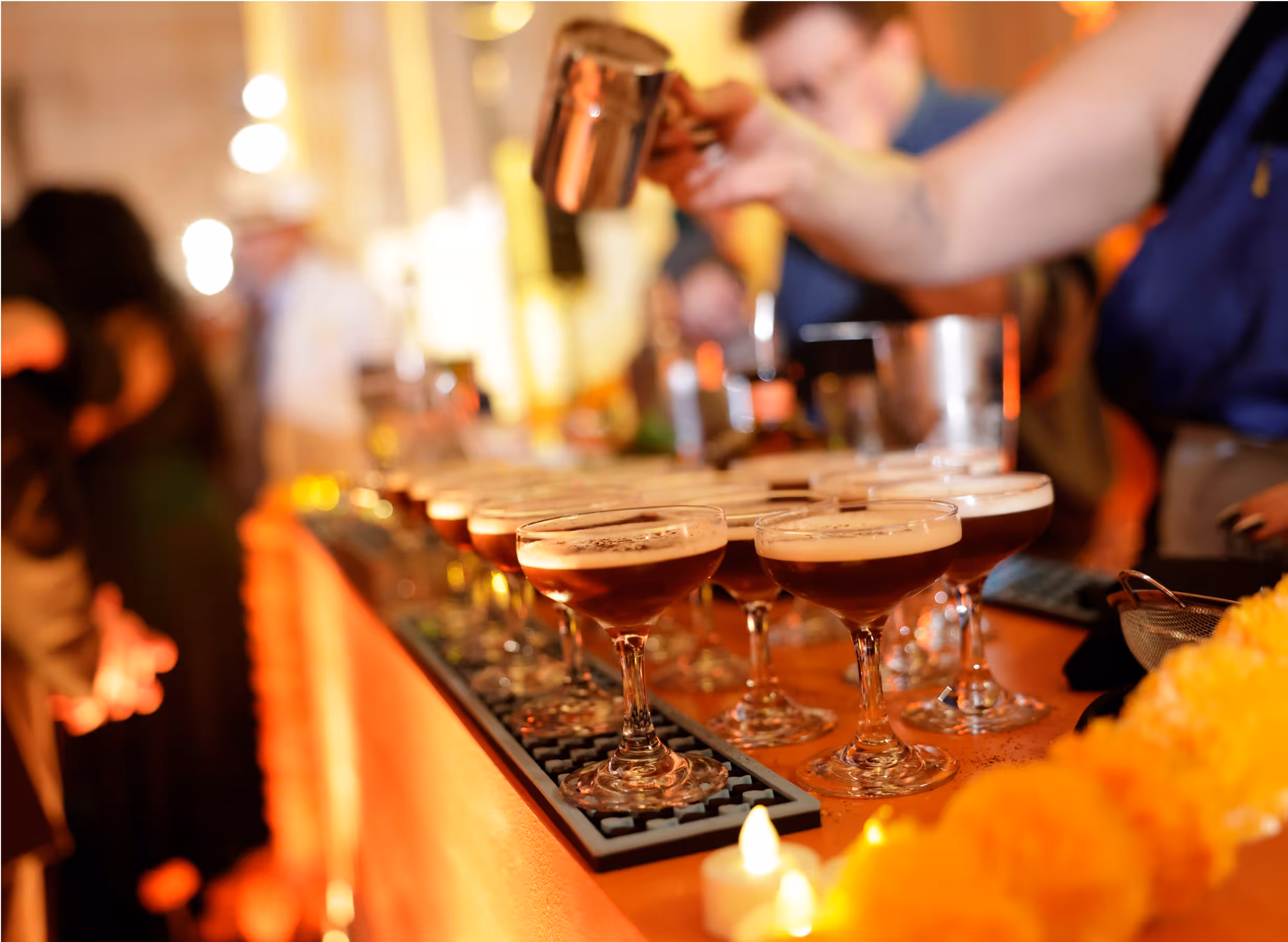 A bartender pours drinks into coupe glasses lined up on a bar, surrounded by warm lighting and blurred people in the background, creating a lively and festive atmosphere.