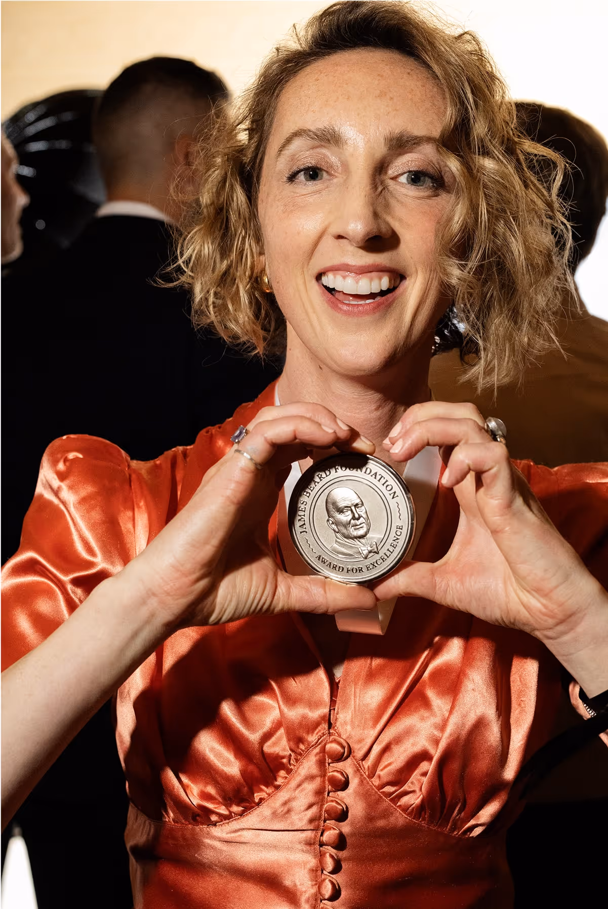 A smiling woman in a shiny red dress holds up a round silver medallion with a face engraved on it. Several people in formal attire stand blurred in the background.