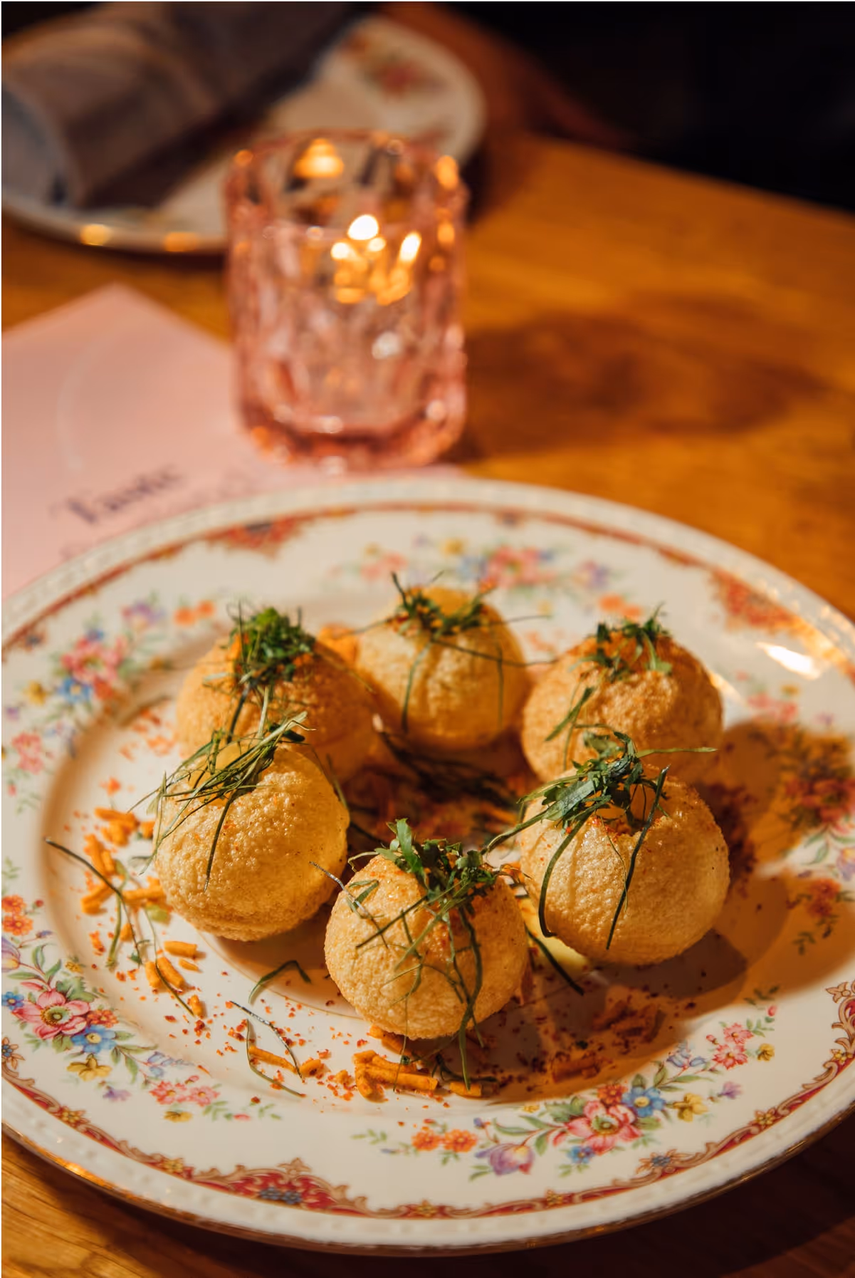 Six round, crispy snacks garnished with herbs are served on a decorative floral plate. A pink glass with a lit candle and a napkin on a plate are visible in the background on a wooden table.