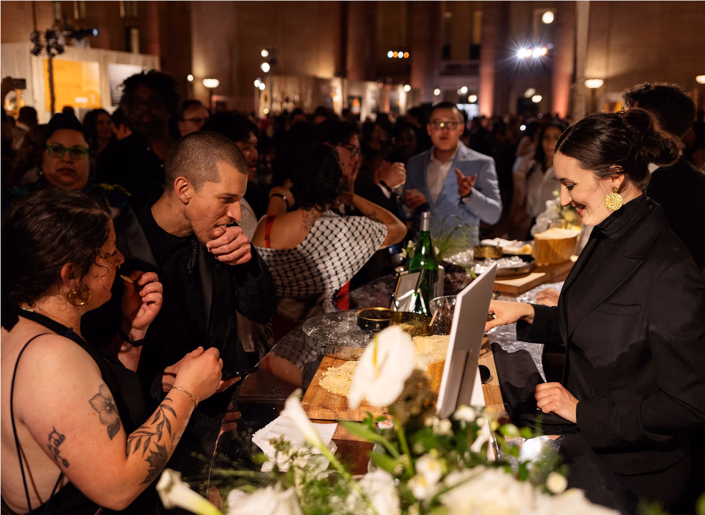 A crowd of people in formal wear gather around a counter at a lively indoor event, where a woman in dark clothing serves food. There are flower arrangements and plates of cheese on the counter.
