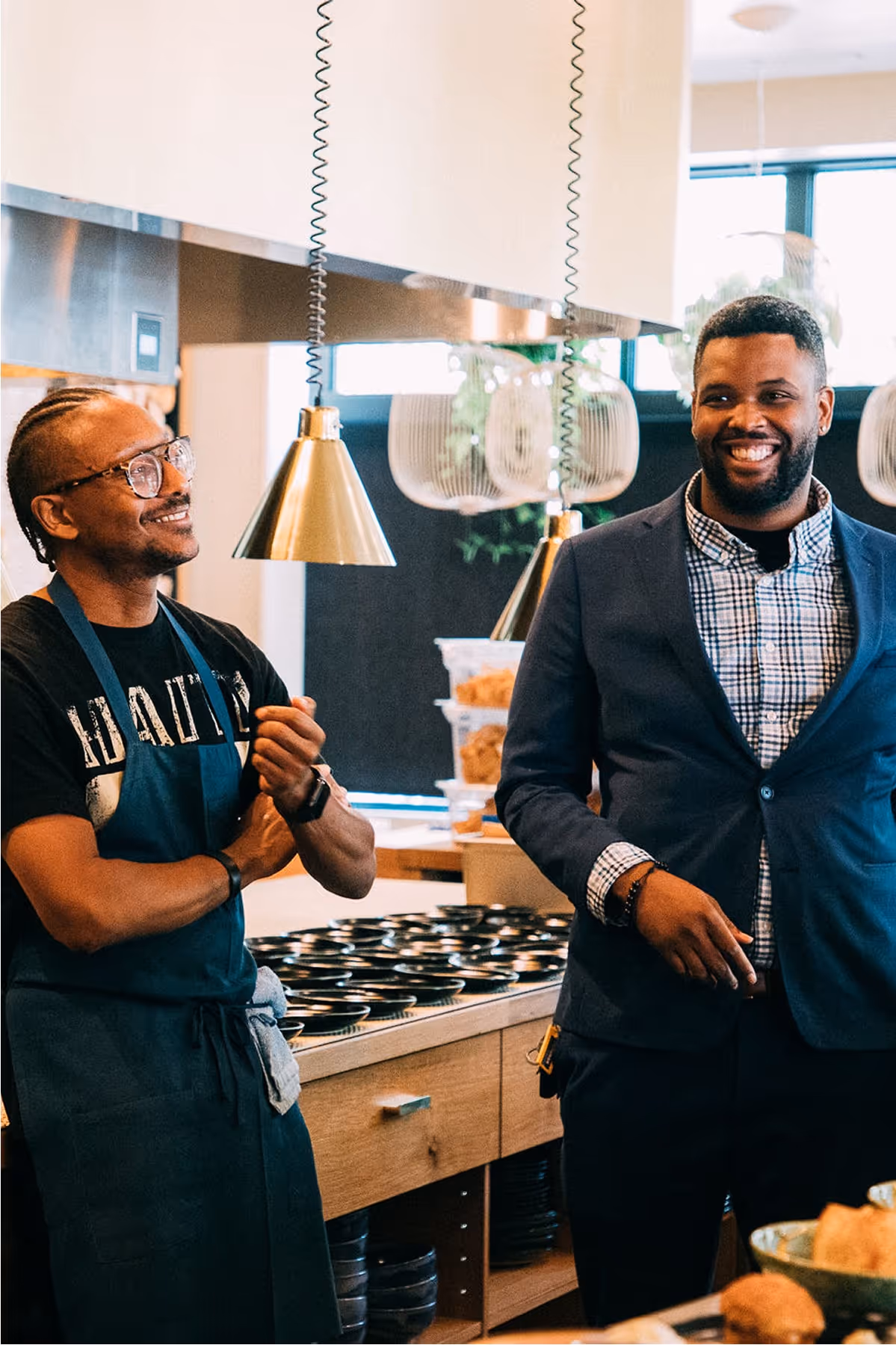 Two men stand and smile in a modern kitchen. One wears glasses, a T-shirt, and an apron, while the other is dressed in a suit. They appear to be enjoying a light-hearted conversation.