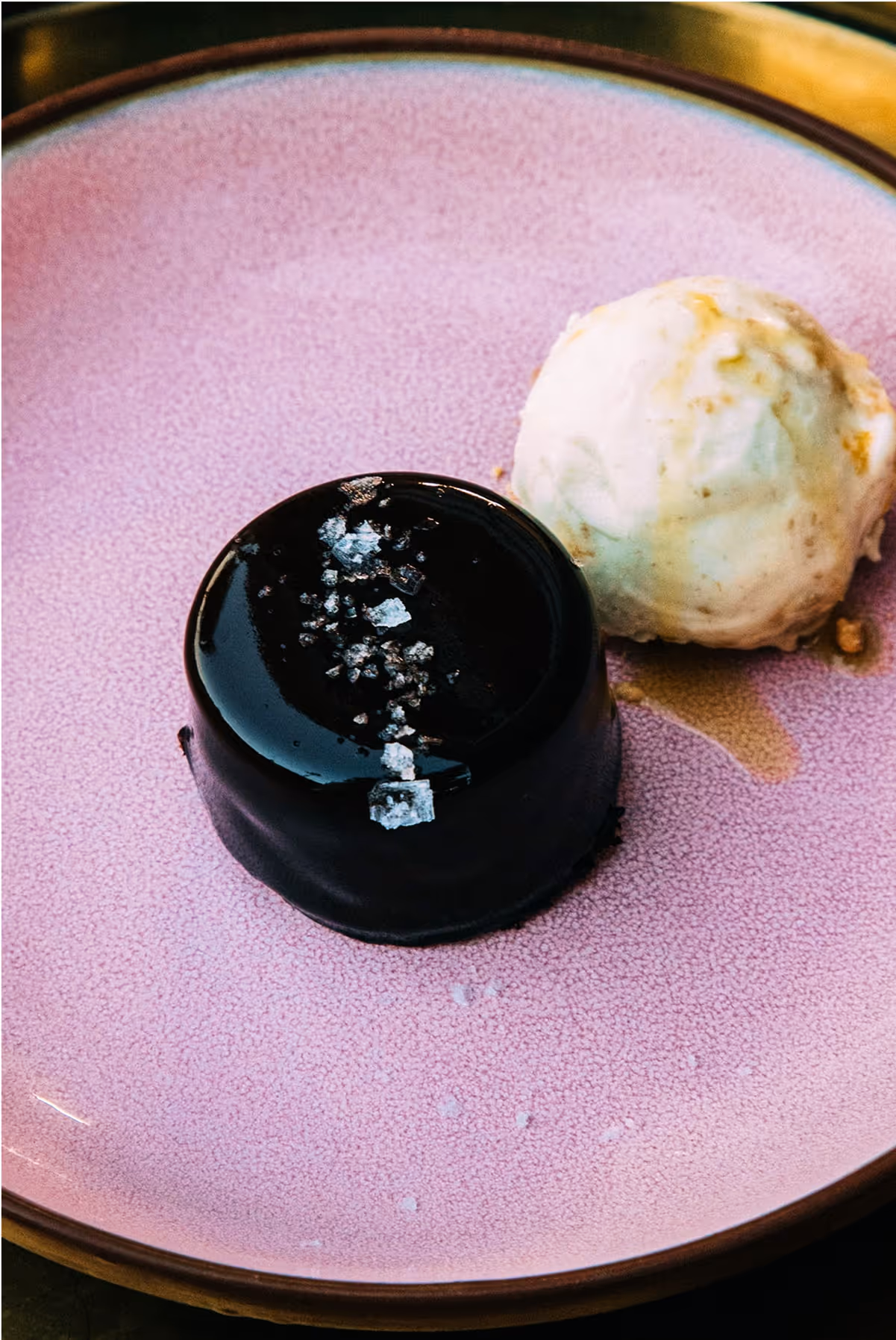 A glossy chocolate dessert topped with coarse sea salt sits on a pink plate beside a scoop of vanilla ice cream.