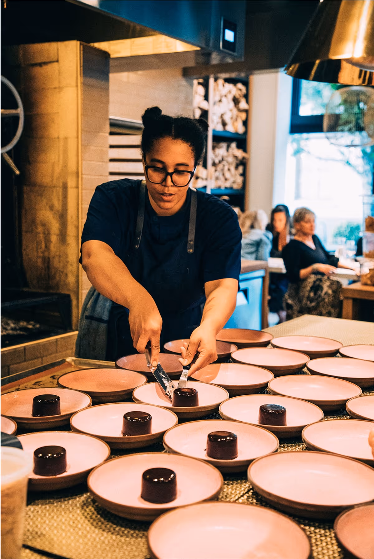 A chef in a blue shirt and glasses carefully places chocolate desserts on pink plates in a busy, modern restaurant kitchen, with several people seated in the background.