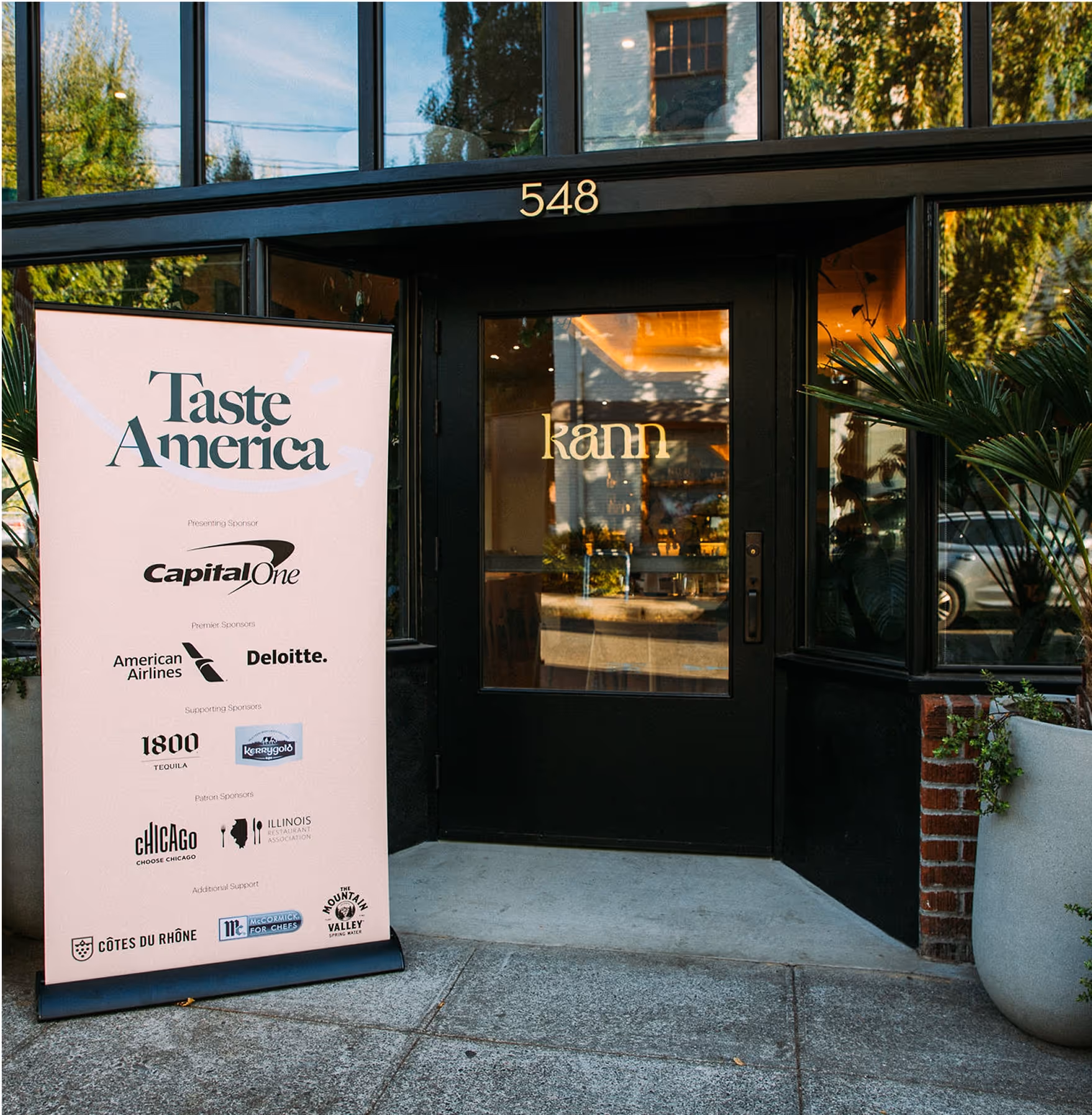 A black-framed glass door entrance to a building labeled Kann with a large peach-colored Taste America event sign listing sponsors like Capital One, American Airlines, and Deloitte, surrounded by potted plants.