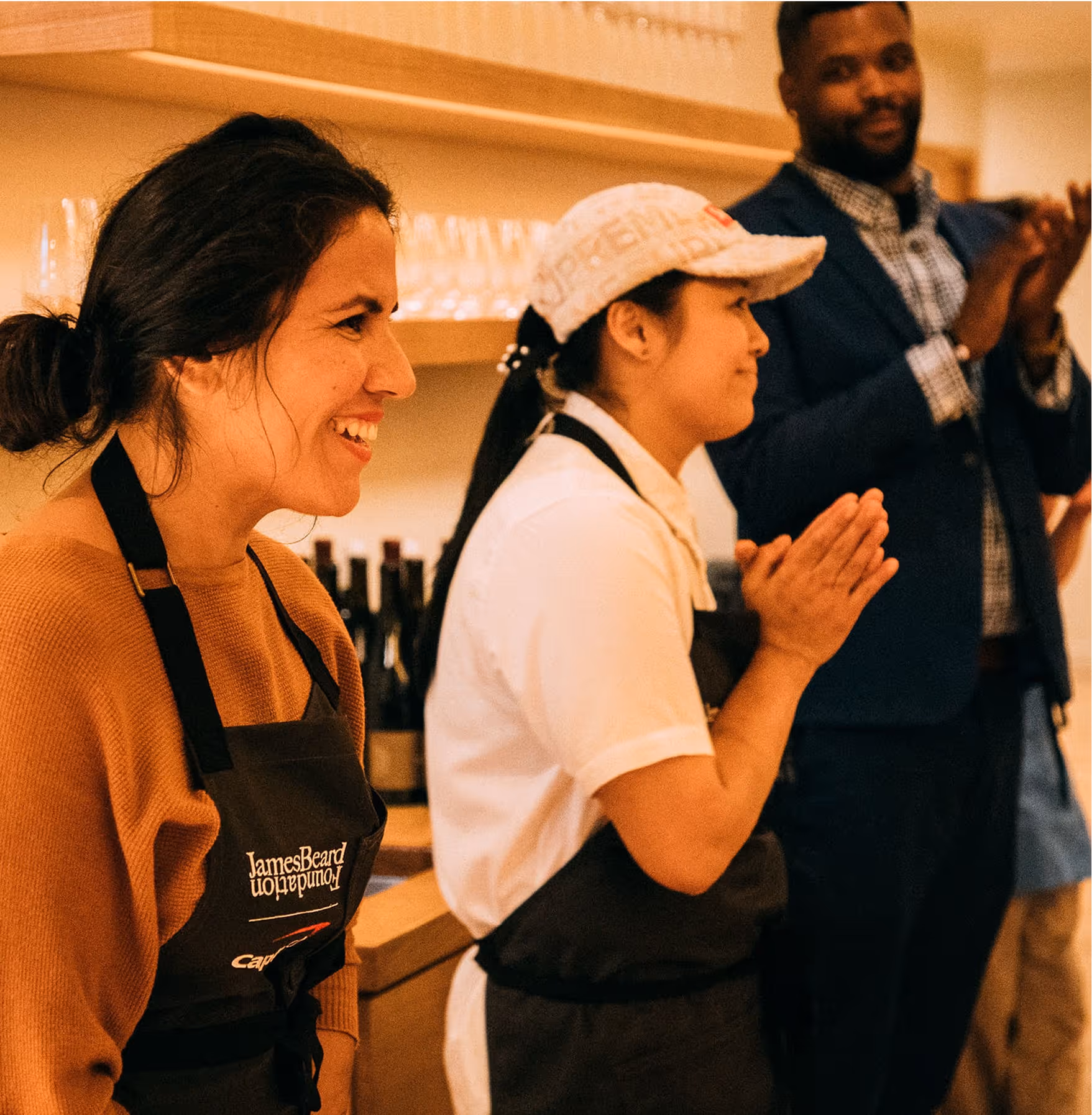 Three people stand indoors clapping and smiling; two women in aprons are in the foreground, and a man in a suit is in the background. Shelves with glasses and bottles are visible behind them.