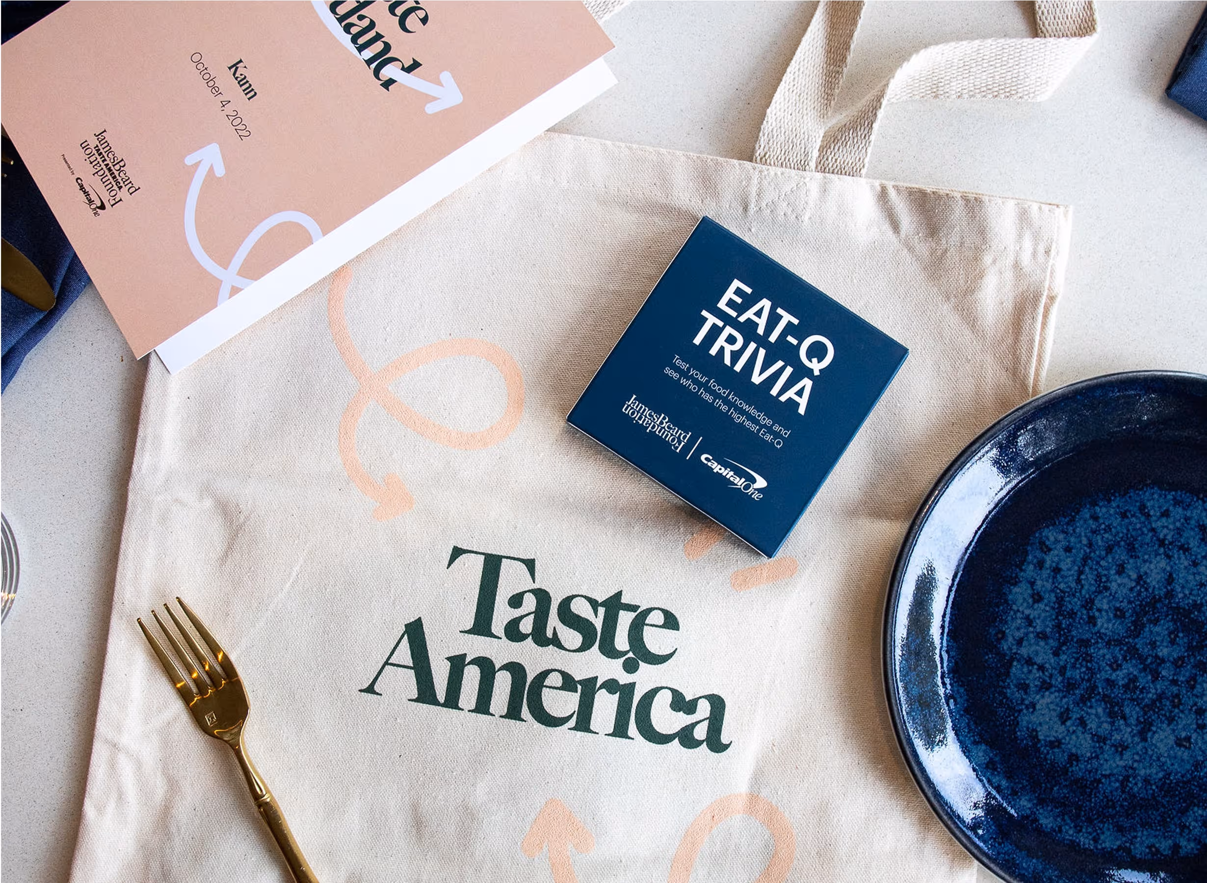 A beige tote bag with “Taste America” printed on it sits on a table alongside a blue plate, a gold fork, an event booklet, and a blue card that reads EAT-Q TRIVIA.