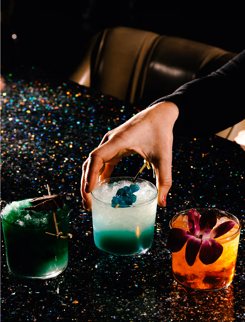 A hand reaches for a blue cocktail with ice on a glittery black table, next to two other colorful drinks—one green with a garnish and one orange with a purple flower.