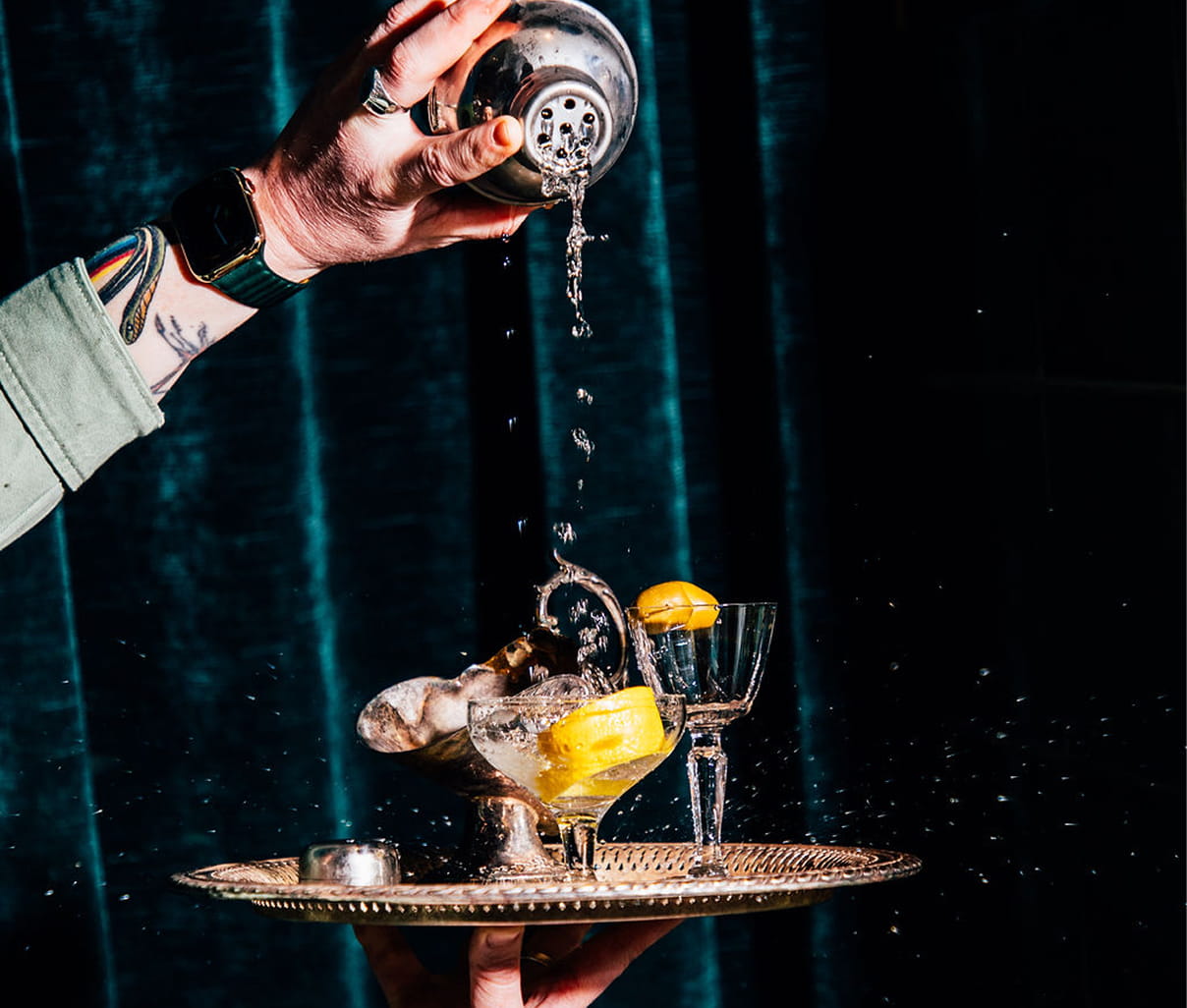 A hand pours a cocktail from a cocktail shaker onto a silver tray filed with multiple coupe glasses in front of a green velvet backdrop.