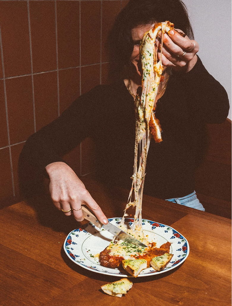 A person pulls a cheesy, saucy piece from a pasta dish on a plate, with the cheese stretching high. 