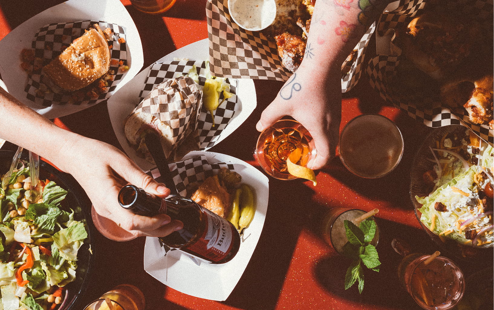 Overhead view of hamburgers, sandwiches, beers, cocktails and sandwiches on a sparkly red table.