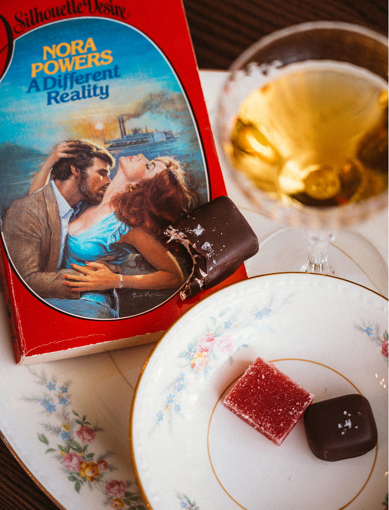 A romance novel titled 'A Different Reality' by Nora Powers with a partially eaten chocolate on it, alongside a glass of amber liquid and a plate with two small confections.