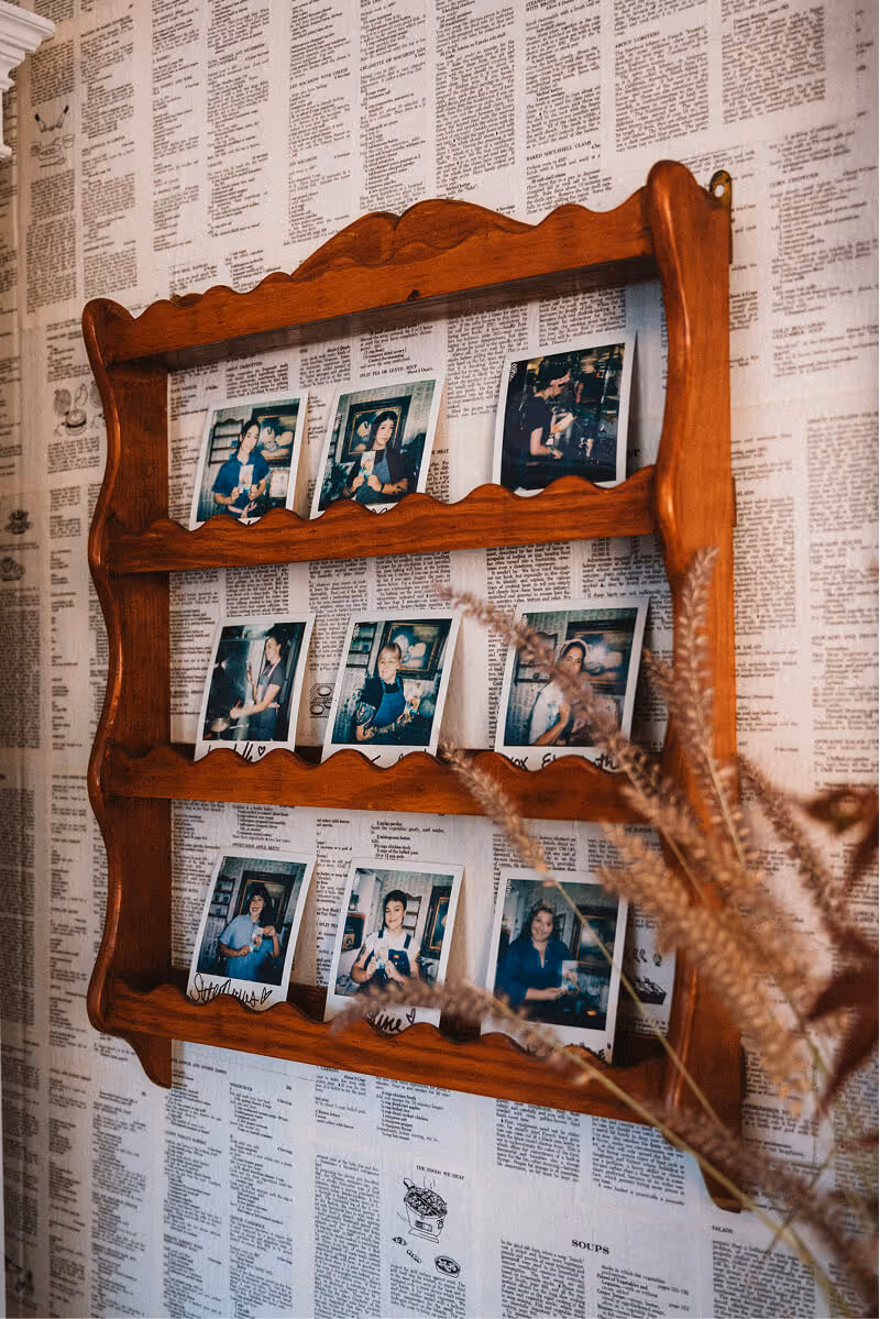 Wooden wall-mounted photo holder with nine Polaroid pictures of people in a cozy setting, against wallpaper covered in newspaper text.