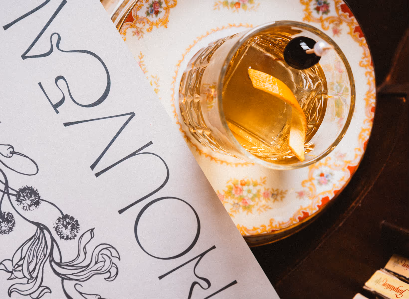 Glass of amber cocktail with a lemon twist and a cherry on a floral patterned saucer next to a section of a printed menu.