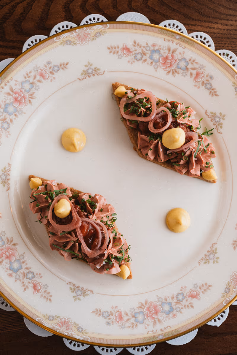 Two triangular toasts topped with pink meat mousse, pickled onions, chives, dollops of mustard, and a red sauce on a floral-patterned plate.