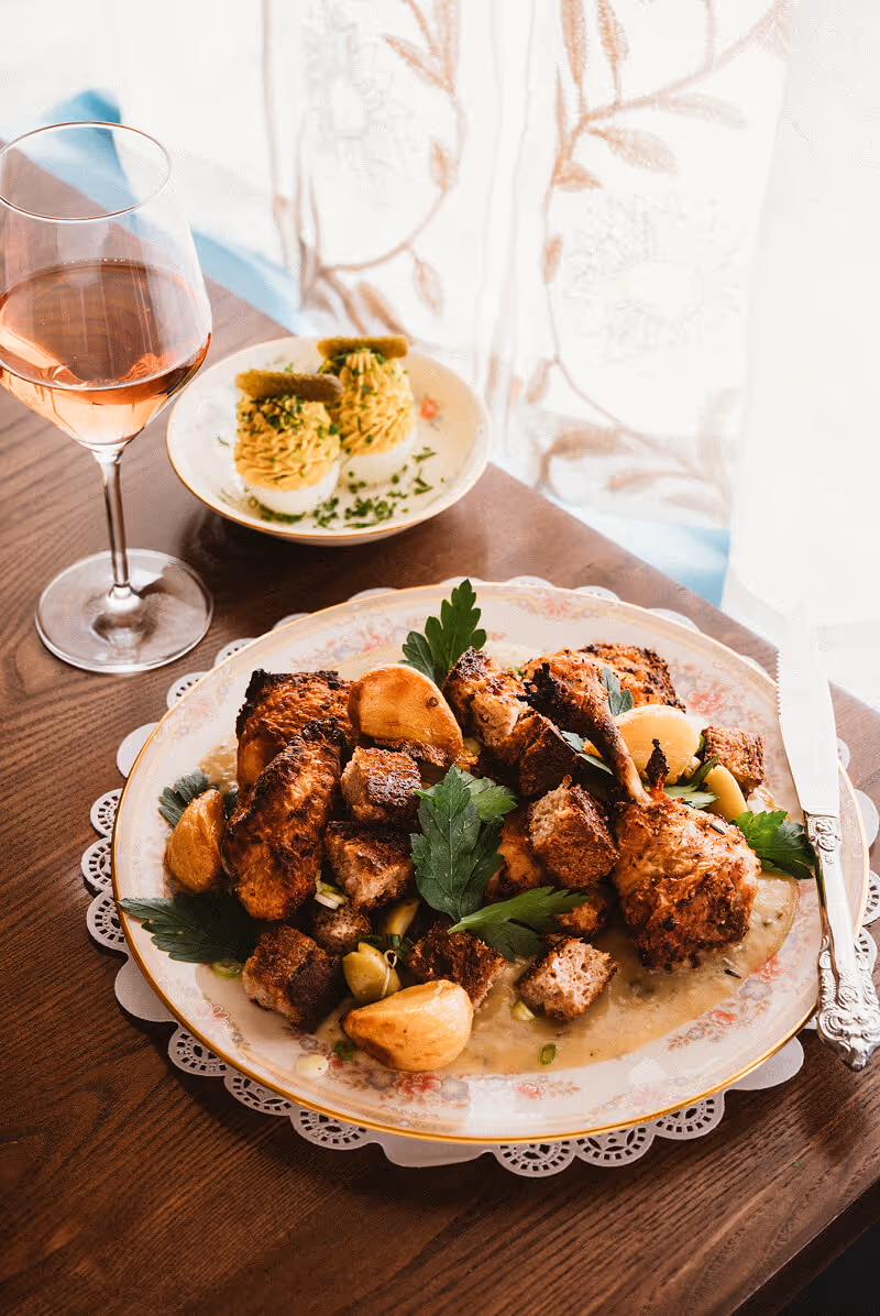 Plate with roasted chicken drumsticks, croutons, garlic cloves, and parsley on a wooden table, served with a small plate of deviled eggs and a glass of rosé wine.
