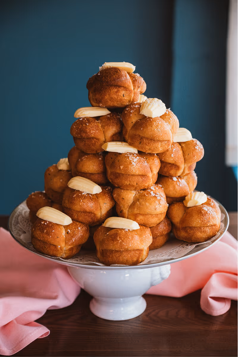 Stack of soft pretzel knots topped with pats of butter and coarse salt on a white pedestal plate with pink cloth on a wooden table.