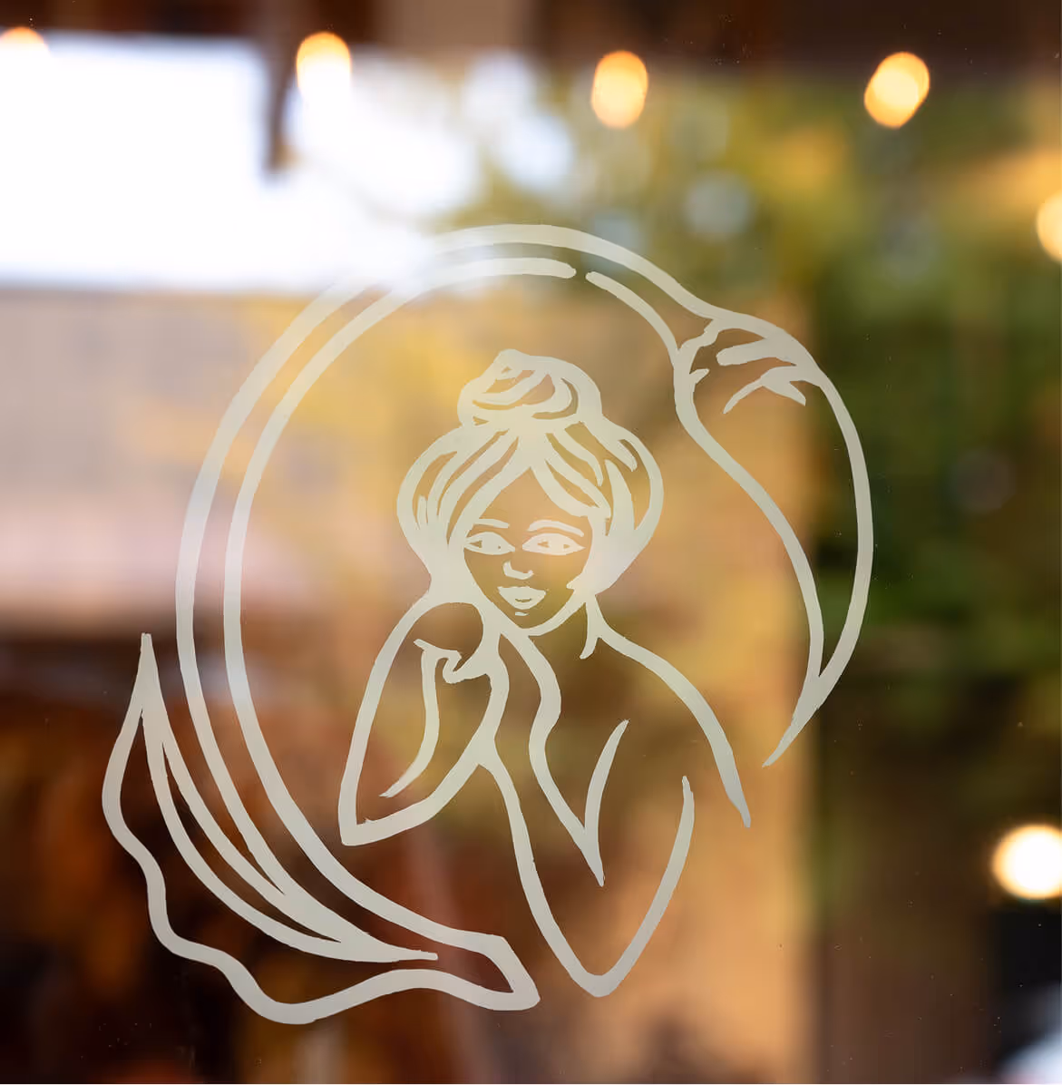 White line art of a woman with hair in a bun, framed by a stylized crescent moon, on a glass surface with blurred background.