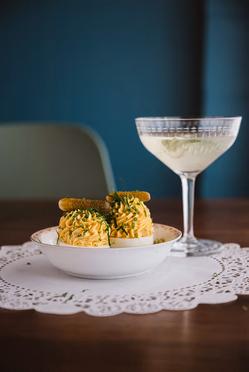 Two deviled eggs garnished with chopped herbs and small pickles on a white plate, next to a clear cocktail glass with a light-colored drink.