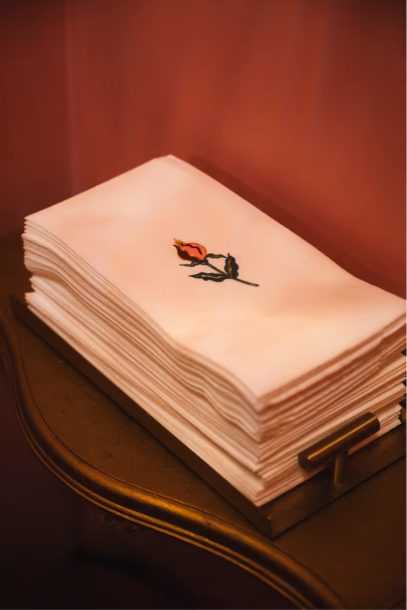 Stack of white napkins embroidered with a red rose laid on a wooden tray on a dark wooden surface.