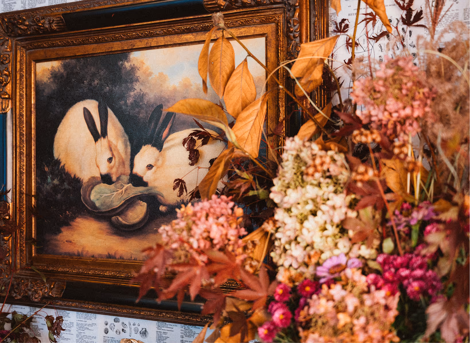 Framed painting of two white rabbits with black ears eating a leafy vegetable, surrounded by autumnal dried flowers and leaves.