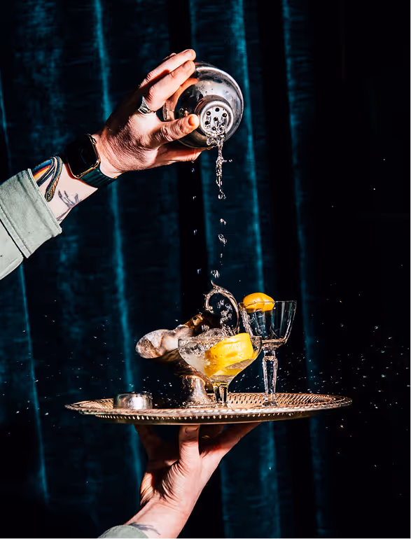 A person pouring liquid from a shaker into a cocktail glass with lemon slices on a brass tray against a dark blue background.
