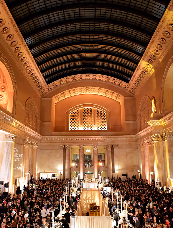 Large crowd gathered inside an ornately decorated hall with a high arched ceiling and lit windows, attending an event with booths and displays.