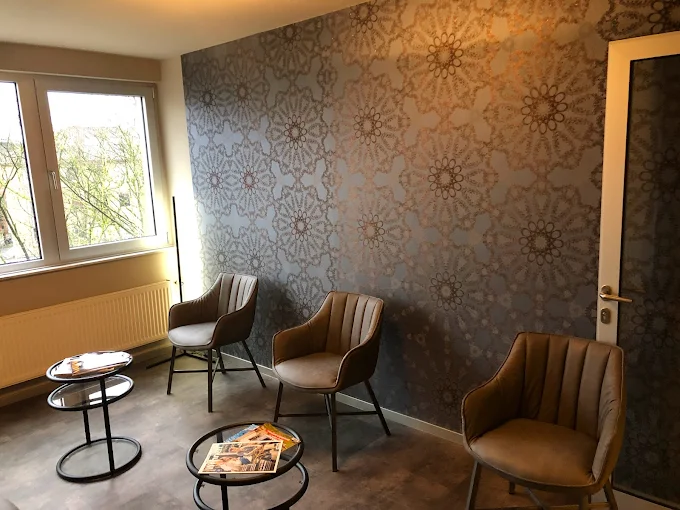 Modern waiting area with three cushioned chairs, two round black metal tables with magazines, and patterned wallpaper near a window and door.