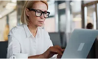 Woman with glasses working on a laptop in a modern office setting.