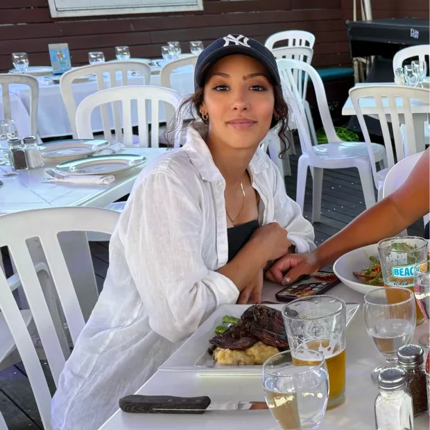 Woman wearing a black cap and white shirt sitting at a table with a plate of food and holding hands with someone next to her.