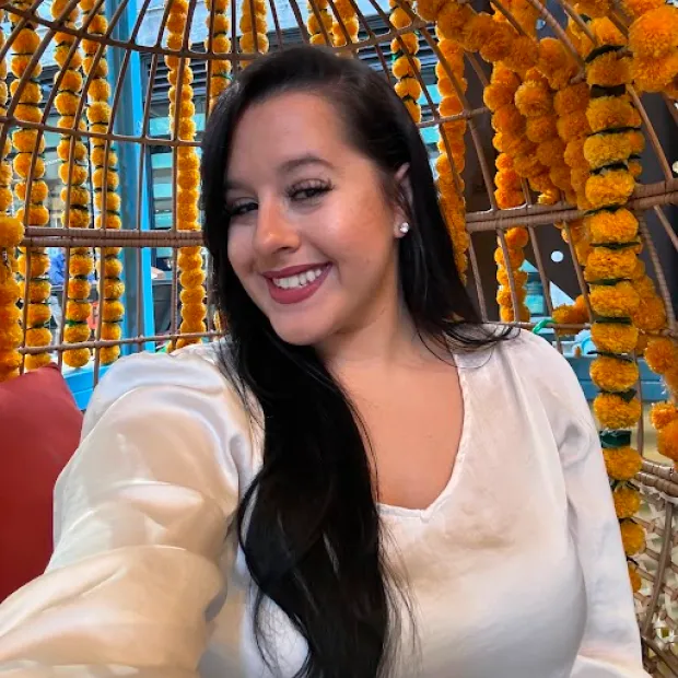 Smiling woman with long dark hair wearing a white blouse, sitting in a wicker chair decorated with orange marigold garlands.