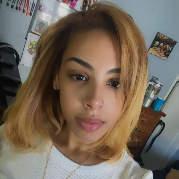 Close-up of a woman with shoulder-length blonde hair wearing a white top, with a nose ring and a neutral expression.