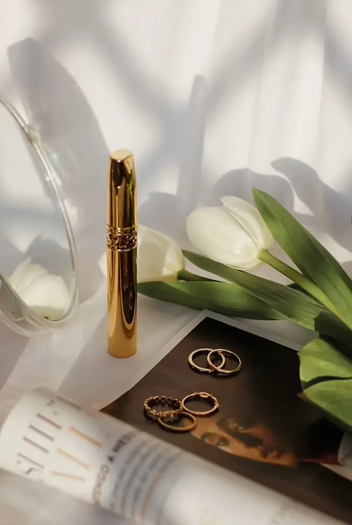 Still life with gold lipstick, white tulips, gold earrings on a photo, and a blurred skincare bottle in soft natural light.