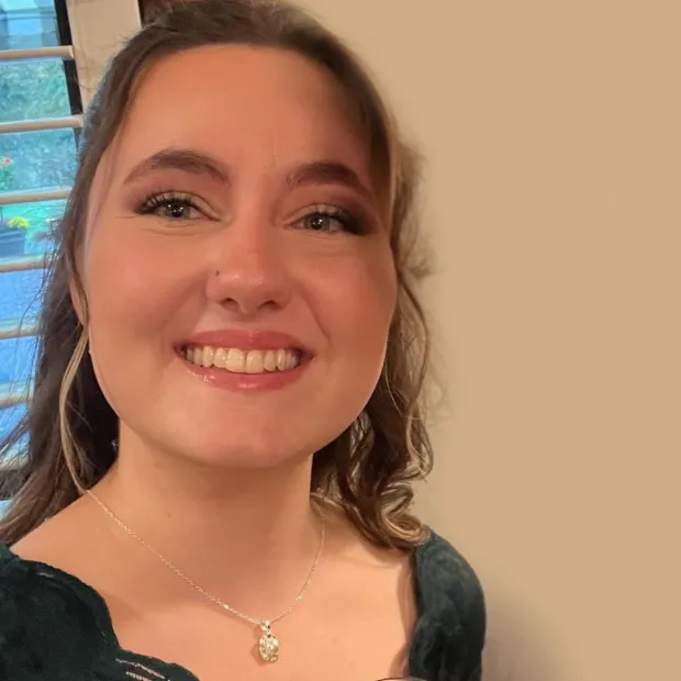Smiling woman with wavy brown hair, wearing a dark green top and a silver pendant necklace.