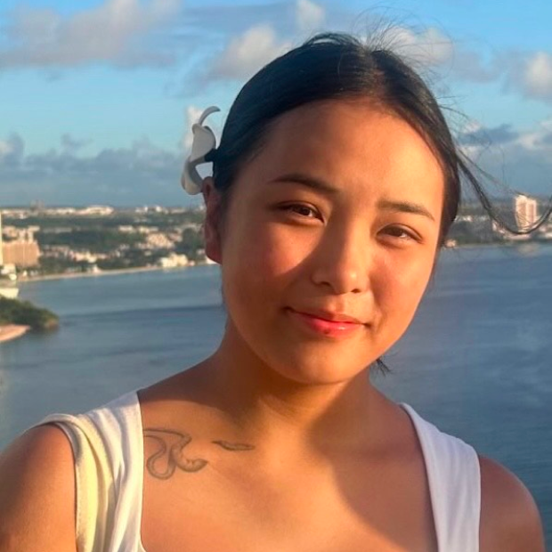 Young woman with a white flower in her hair, smiling softly with a waterfront cityscape in the background.