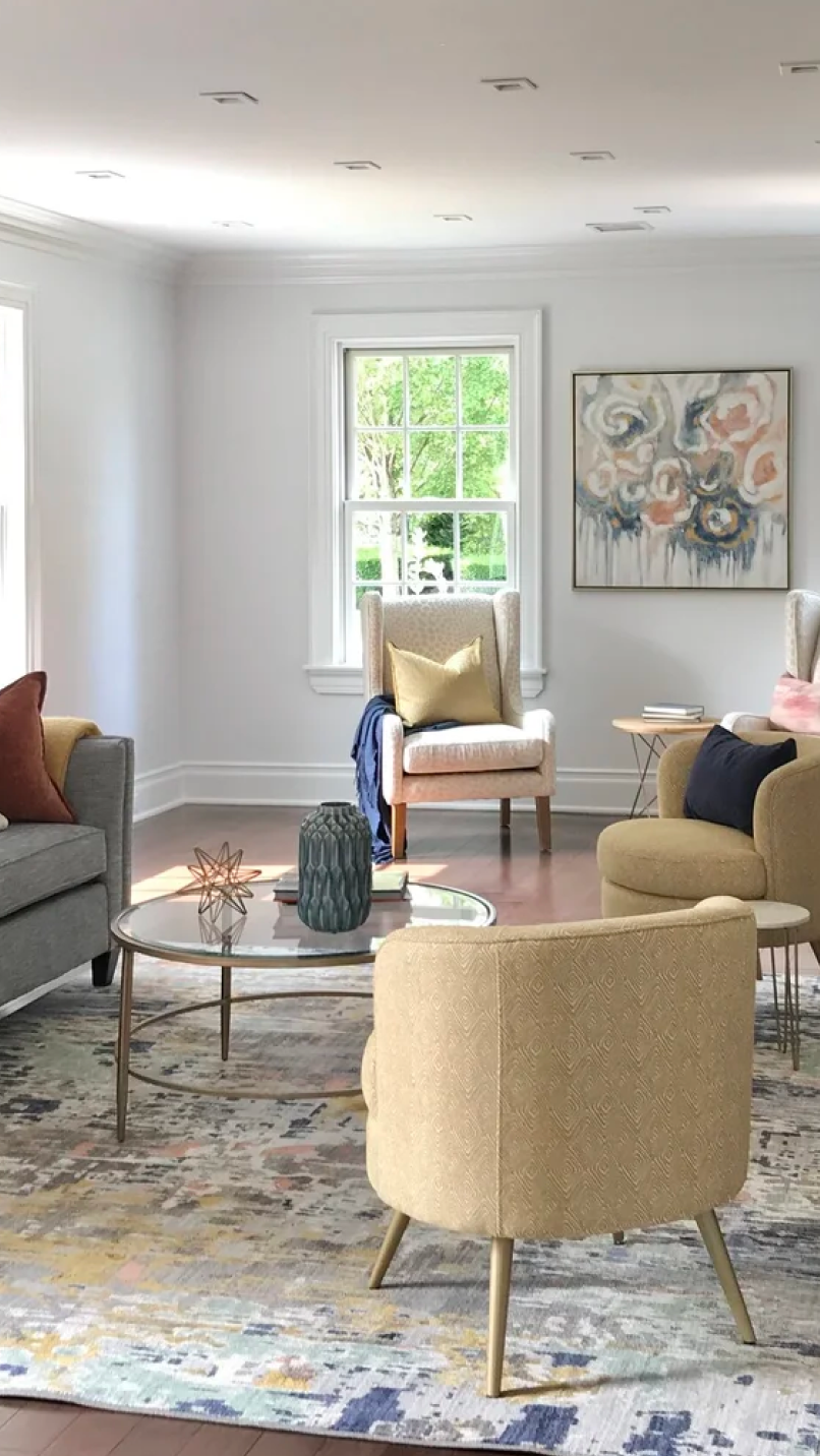 Bright living room with a gray sofa, two beige armchairs, a round glass coffee table, and abstract art on the wall.