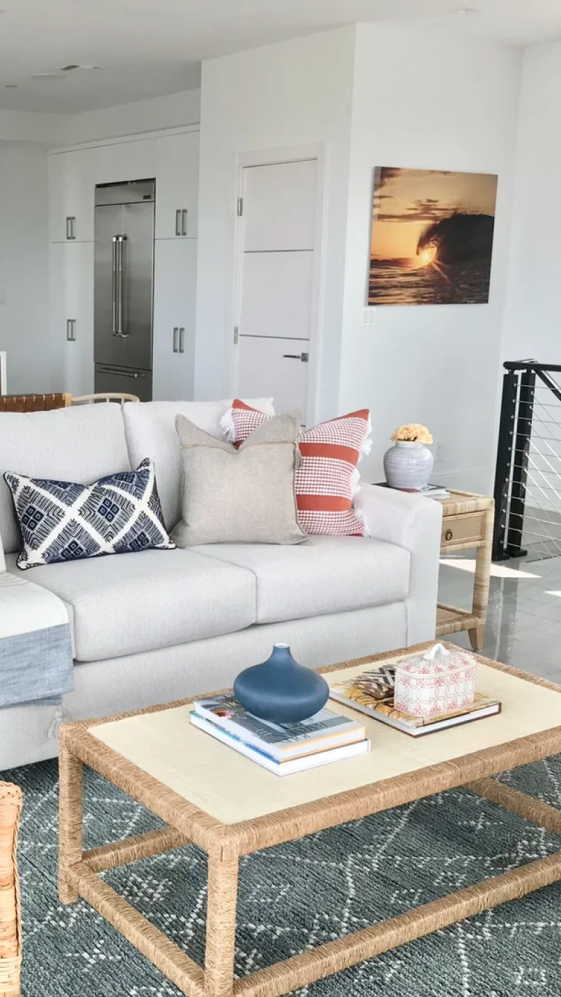 Modern living room with light gray sofa, patterned throw pillows, wicker coffee table with books and blue vase, and ocean wave sunset photo on wall.