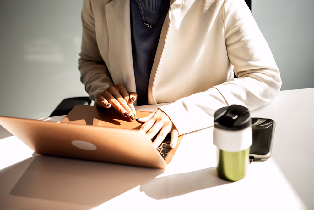 Person in a light blazer typing on a laptop at a white desk with a green and white travel mug and a smartphone.