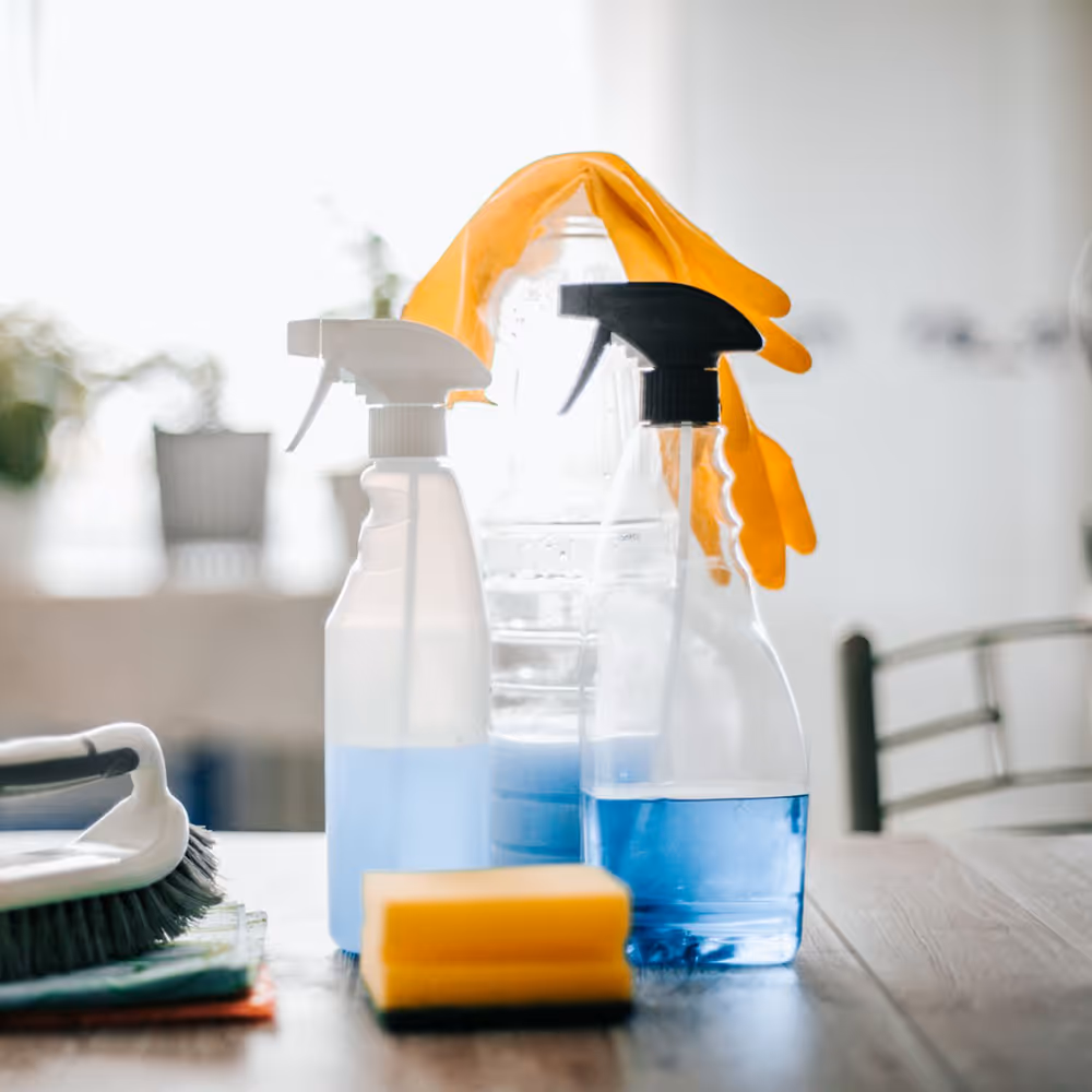Cleaning supplies on a wooden table including spray bottles with blue liquid, a sponge, a brush, and yellow rubber gloves draped over a water bottle.