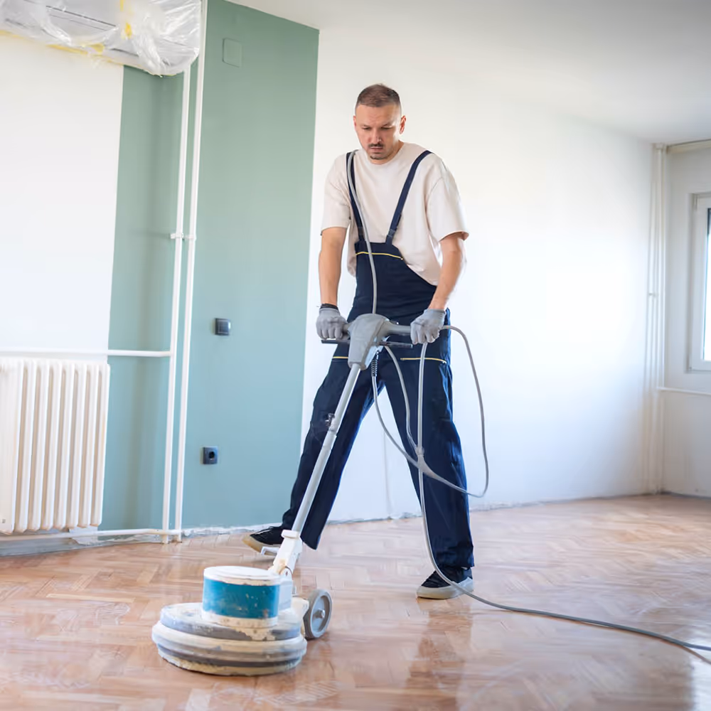 Man in overalls polishing a wooden parquet floor with a floor polishing machine in a bright room.