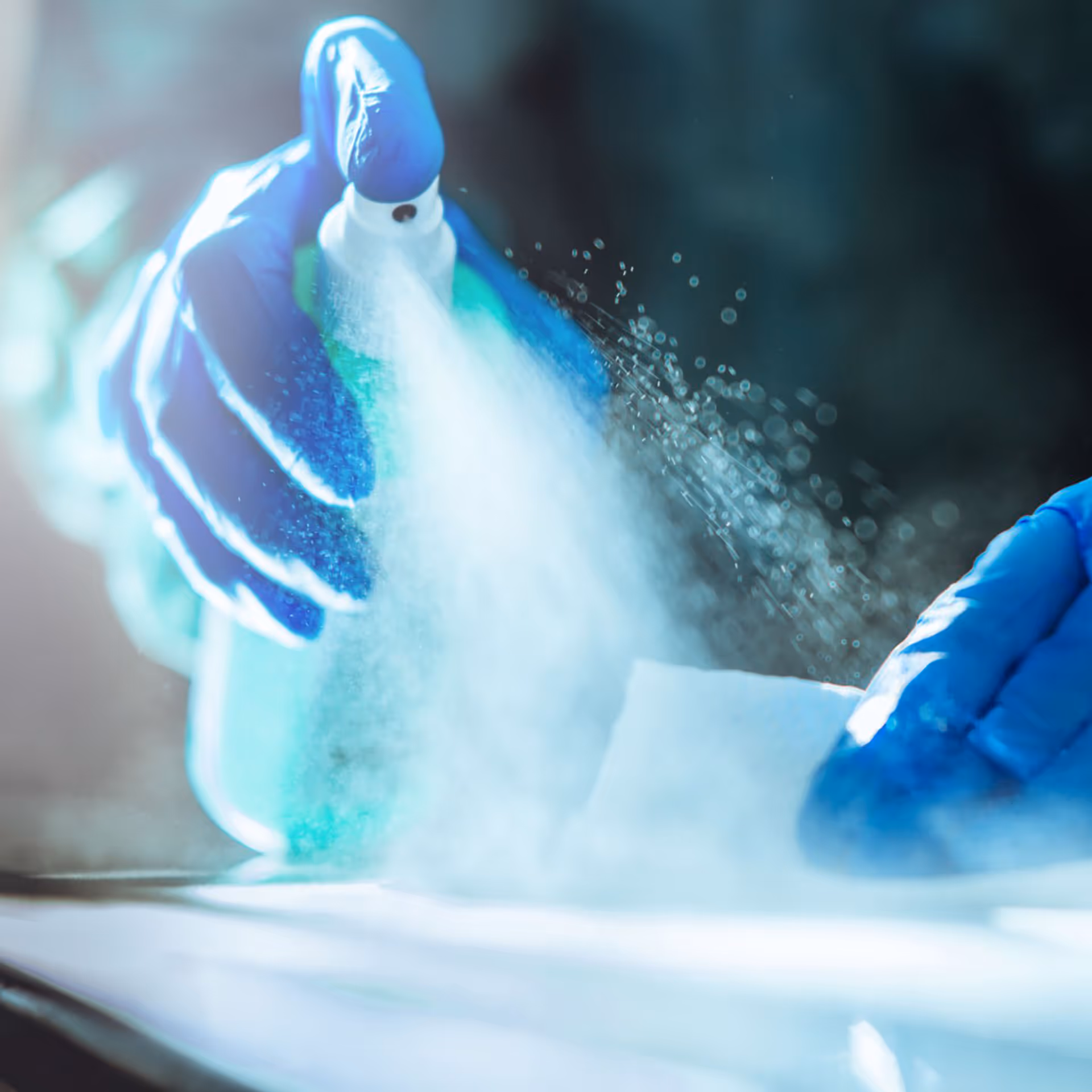 Person wearing blue gloves spraying cleaning solution onto a surface with a white cloth.