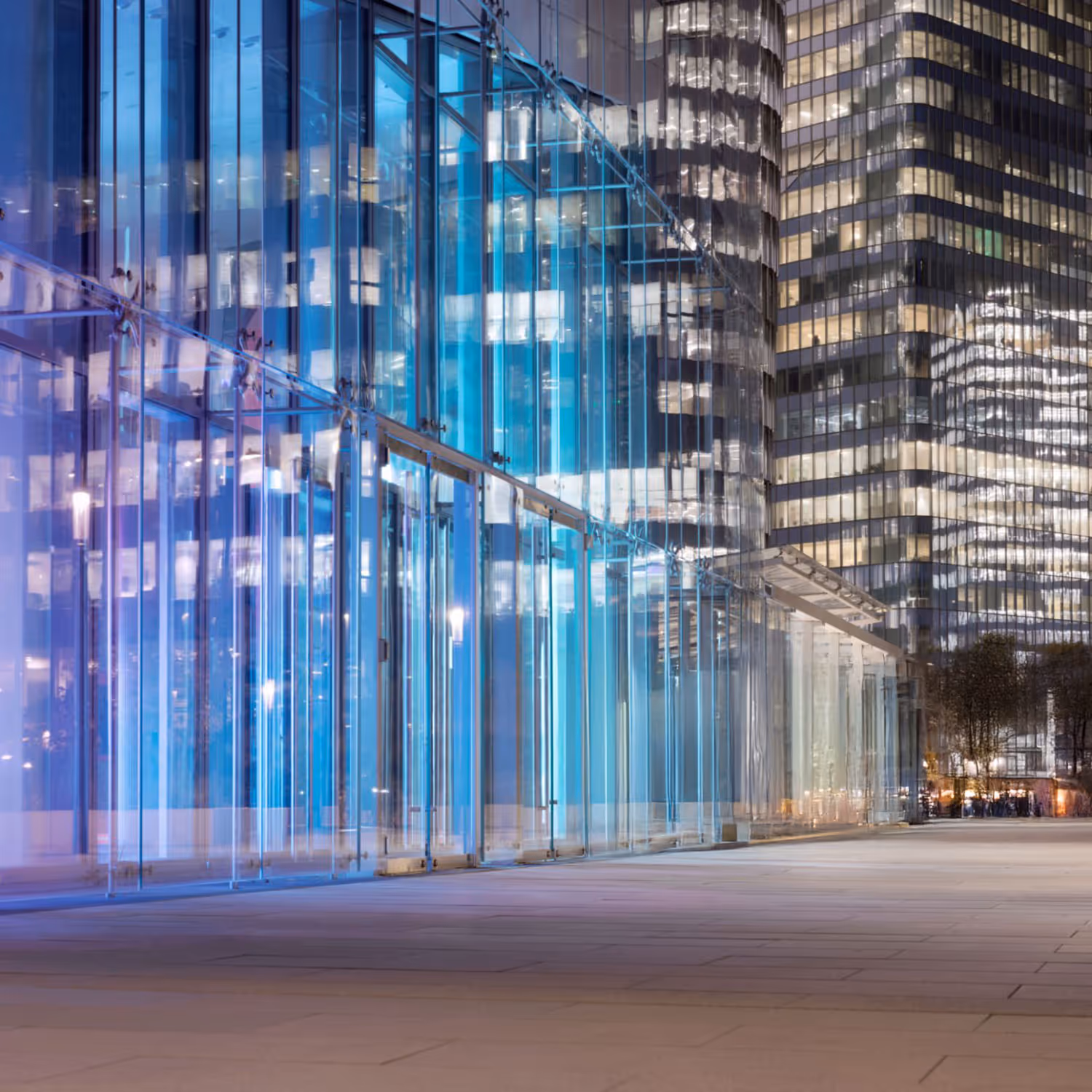 Glass building facade illuminated with blue light reflecting nearby skyscrapers at night.